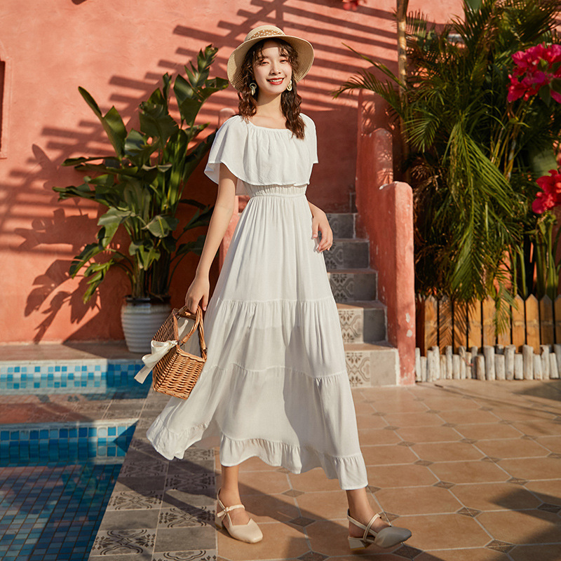 Off-shoulder frill maxi dress with summer-ready design, woman in white dress and hat by a pool.